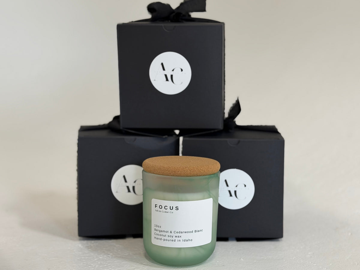 A frosted glass jar Focus candle with a cork lid stands in front of three black gift boxes with black ribbons and white circular logos. Bergamot & Cedarwood Blanc